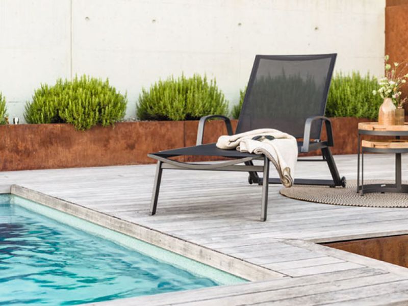 Ein moderner Liegestuhl mit schwarzer Bespannung steht an einem Pool auf einer Holzterrasse, daneben ein kleiner runder Beistelltisch aus Holz mit einem Blumenarrangement obenauf und einem gefalteten Tuch darunter. Das Ensemble ist umgeben von einer dezenten Gartenlandschaft mit sauber geschnittenen Büschen und einer rostfarbenen Wand im Hintergrund.