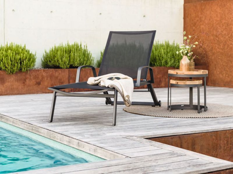 Ein moderner Liegestuhl mit schwarzer Bespannung steht an einem Pool auf einer Holzterrasse, daneben ein kleiner runder Beistelltisch aus Holz mit einem Blumenarrangement obenauf und einem gefalteten Tuch darunter. Das Ensemble ist umgeben von einer dezenten Gartenlandschaft mit sauber geschnittenen Büschen und einer rostfarbenen Wand im Hintergrund.