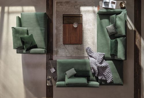 Polstergruppe in sattem Grün, aus der Vogelperspektive fotografiert. Zentrale Anordnung um einen Holz-Couchtisch auf beigem Teppich. Kissen und gestrickte Decke sorgen für Gemütlichkeit. Licht- und Schatteneffekte verleihen Tiefe und Wärme.