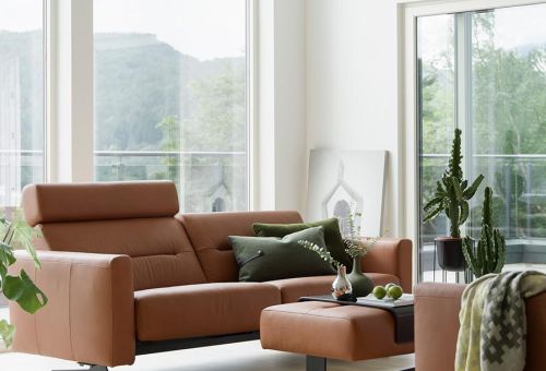 Das Bild zeigt ein modernes Wohnzimmer mit großen Fenstern, die viel natürliches Licht hereinlassen. Im Vordergrund befindet sich ein elegantes Ledersofa in einem warmen Braunton, begleitet von einem passenden Fußhocker. Auf dem Sofa liegen grüne Dekokissen, und ein kleiner Beistelltisch mit einer Blumenvase und einigen dekorativen Gegenständen steht davor. Im Hintergrund sind ein hoher Kaktus, weitere Zimmerpflanzen und ein gerahmtes Bild zu sehen, das lässig an die Wand gelehnt ist. Die helle und minimalistische Einrichtung wird durch die Aussicht ins Freie, die durch die großen Fenster sichtbar ist, ergänzt.
