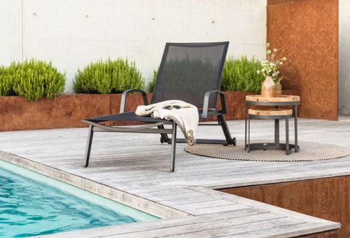Ein moderner Liegestuhl mit schwarzer Bespannung steht an einem Pool auf einer Holzterrasse, daneben ein kleiner runder Beistelltisch aus Holz mit einem Blumenarrangement obenauf und einem gefalteten Tuch darunter. Das Ensemble ist umgeben von einer dezenten Gartenlandschaft mit sauber geschnittenen Büschen und einer rostfarbenen Wand im Hintergrund.