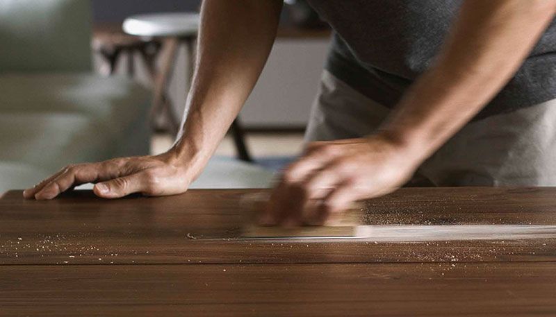 Eine Person reinigt eine Holzoberfläche mit der flachen Hand und etwas Reinigungsmittel, welches auf dem Tisch eine weiße Spur hinterlässt. Der Fokus liegt auf den Händen und dem Tisch, wobei der Hintergrund mit Möbelstücken unscharf ist.