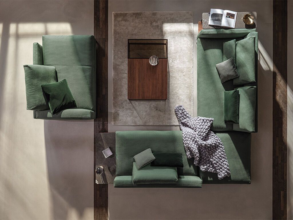 in modernes Wohnzimmer mit einem großen, beigen Ecksofa, auf dem ein Golden Retriever liegt. Das Sofa ist mit mehreren Kissen dekoriert. Im Vordergrund steht ein runder Couchtisch mit Dekorationen. Im Hintergrund sind hohe Fenster mit halbtransparenten Vorhängen zu sehen, sowie eine Stehlampe und ein grüner Tischleuchter auf einem Beistelltisch.