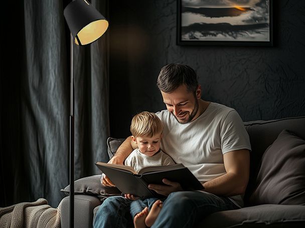 Ein Vater, der seinem Sohn aus einem Buch vorliest. Sie sitzen beide zusammen auf einem Sofa und eine Stehlampe spendet ihnen Licht. 